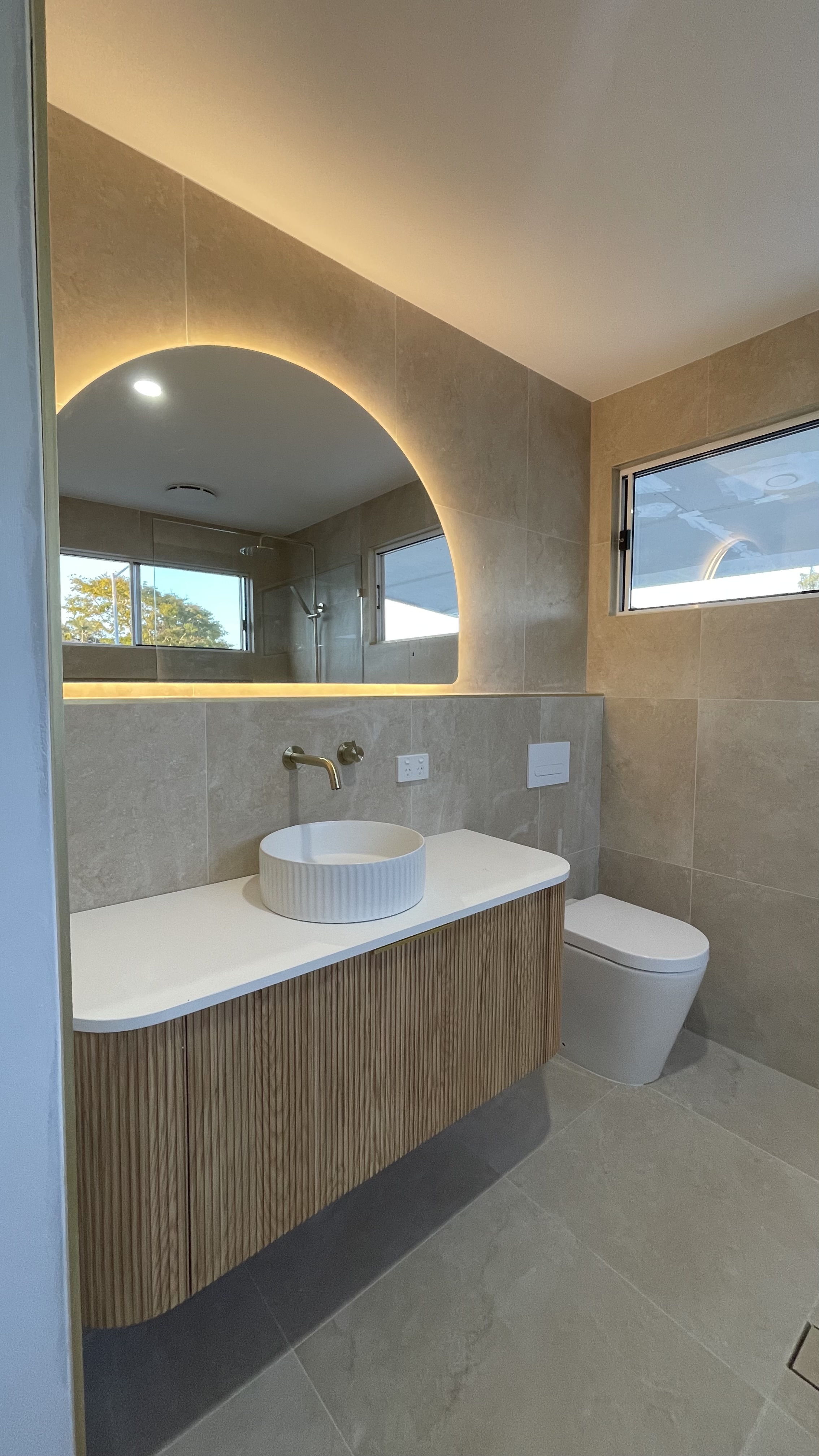 Bathroom with an illuminated curved mirror, fluted timber vanity and warm stone tiles