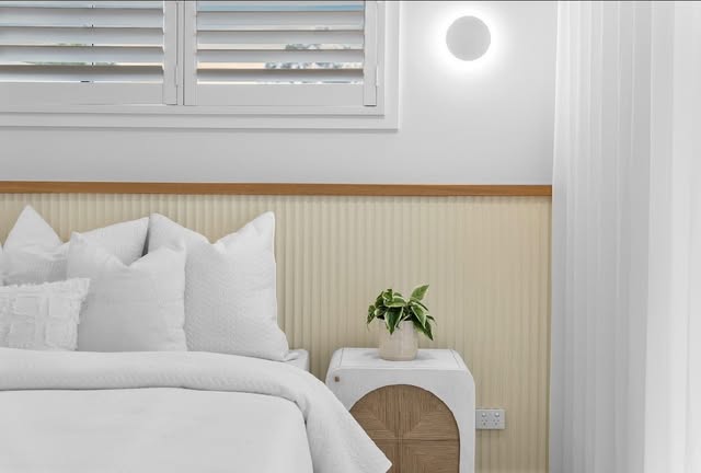 Styled bedroom with fluted wall panelling, timber trim, white bedding and a round wall light